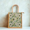 A medium-sized jute bag with white daisy-style floral print on a green background and natural jute handles.
