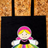 Black fabric tote bag featuring a colorful cartoon-style Mexican doll illustration with traditional dress and long shoulder straps.
