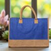 A rectangular tote bag with a vibrant royal blue jute body and a natural brown jute base and handles. The bag is presented against a light wall with a soft-focus background of stacked bags or materials.