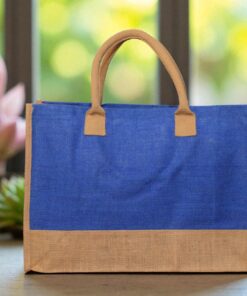 A rectangular tote bag with a vibrant royal blue jute body and a natural brown jute base and handles. The bag is presented against a light wall with a soft-focus background of stacked bags or materials.