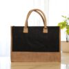 A rectangular tote bag with a black jute body and a natural brown jute base and handles. The bag is presented against a light wall with a soft-focus background of stacked bags or materials.