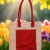 A rectangular natural off-white cotton canvas tote bag featuring a prominent red rectangular pocket on its front and two matching red fabric handles. The bag is hung against a light