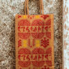 A medium-sized rectangular jute bag featuring a traditional Indian folk art print in red and yellow on a natural jute background.